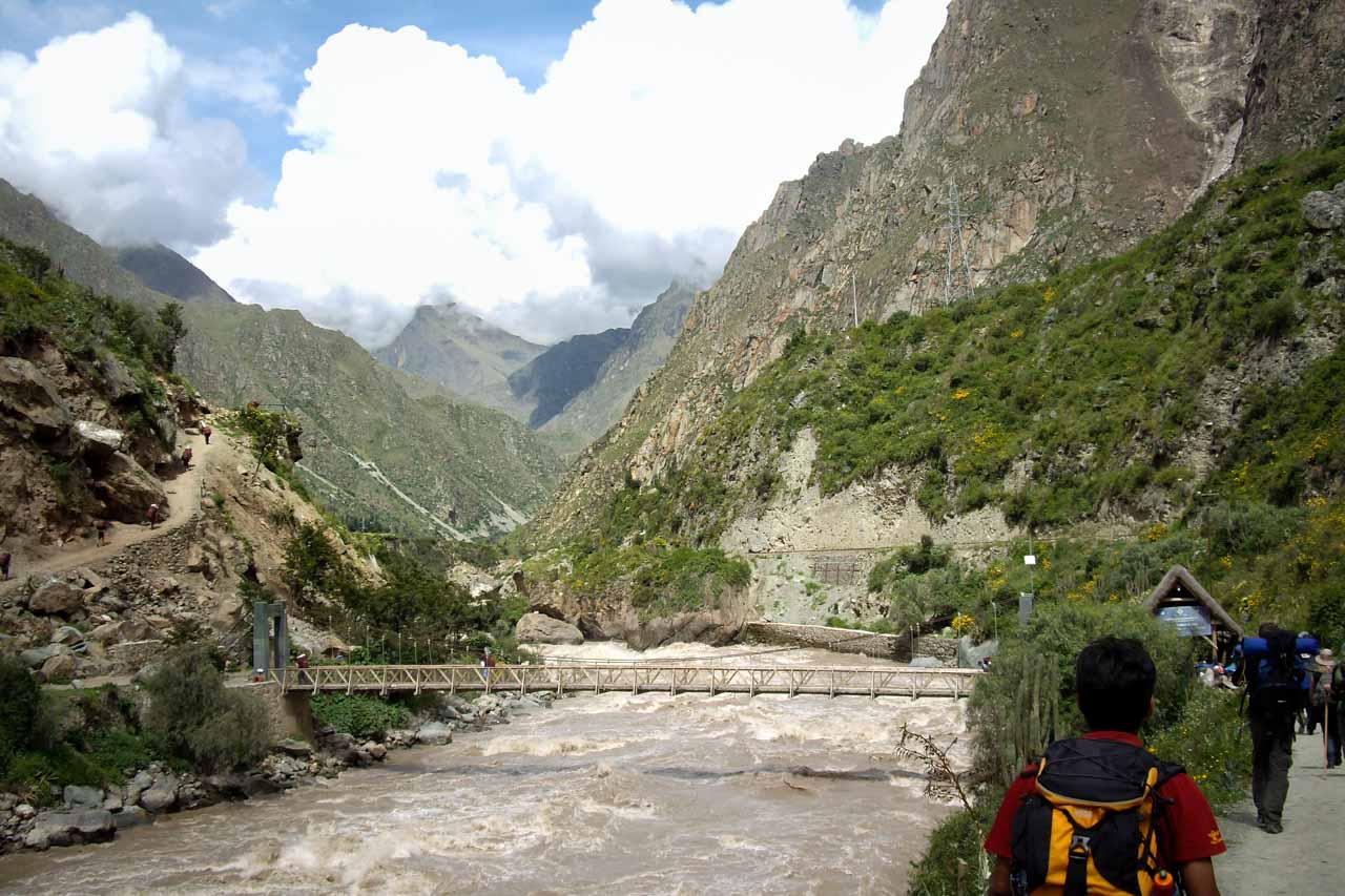 Narrow suspension bridge across muddy river with mountains in the background