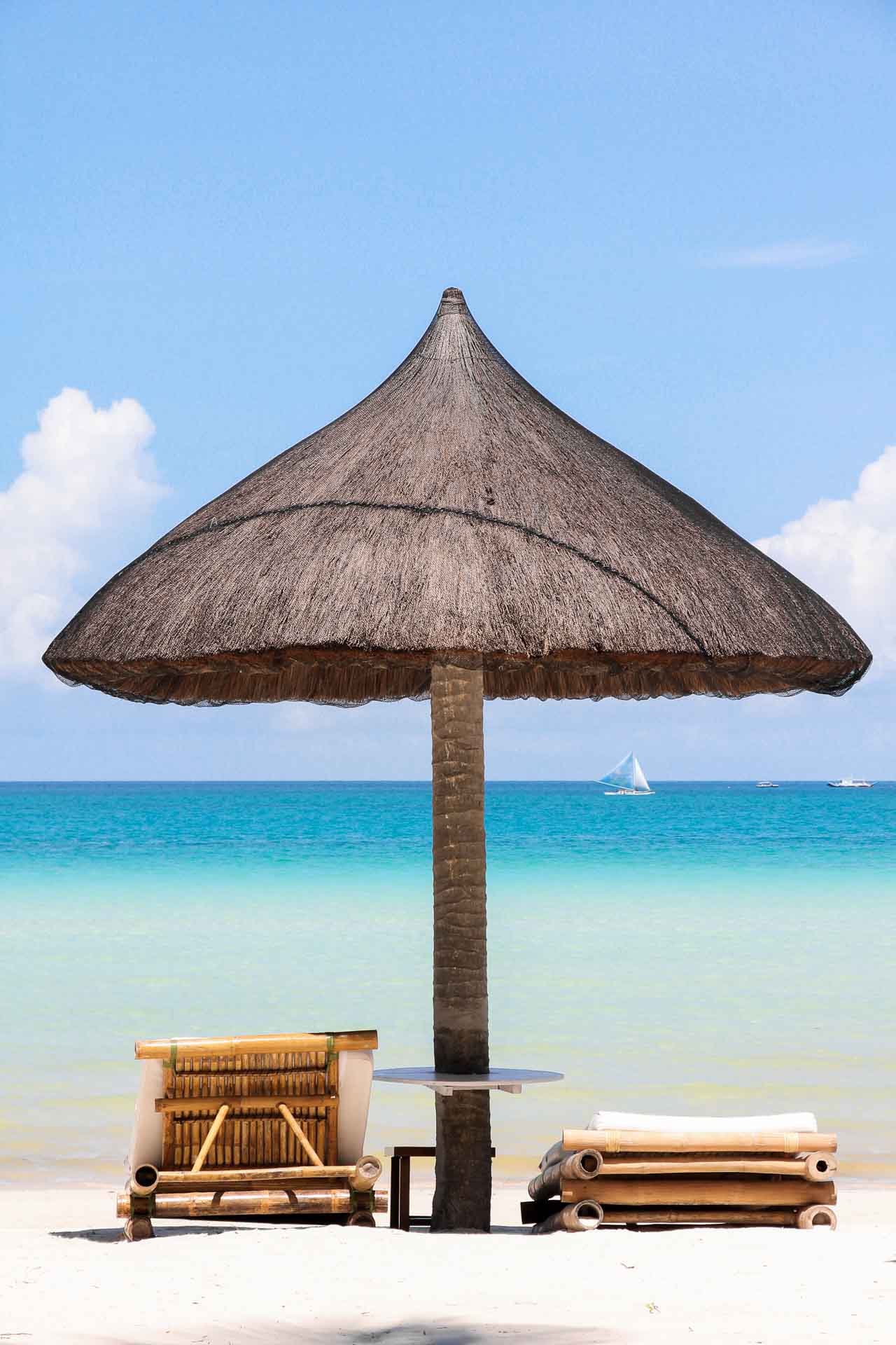 Thatched umbrella and sun loungers on tropical beach with sail boats in background