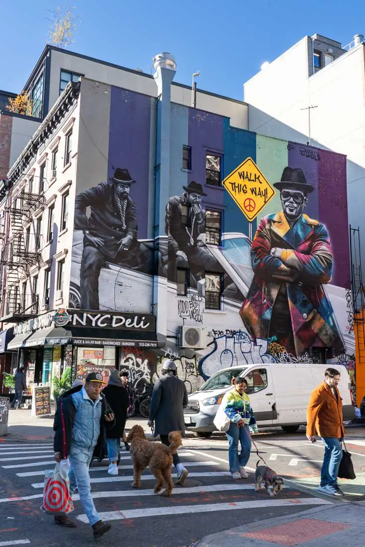 Mural of Run DMC with street sign reading "walk this way" in foreground are people crossing street