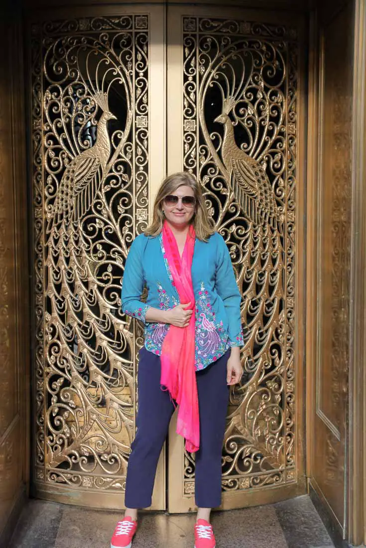 Woman wearing teal and pink kebaya embroidered with peacocks, standing infront of ornate bras doors with peacock motifs