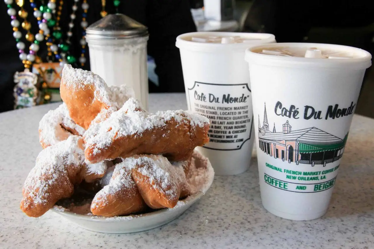 White plate piled high with beignets and two disposable coffee cups reading Cafe Du Monde - Must-Eat New Orleans Foods