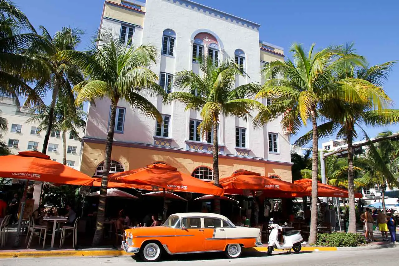Orange and white vintage car parked infront of hotel with palm trees and orange umbrellas