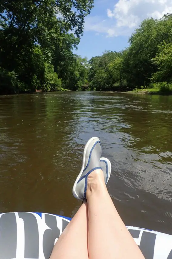 Woman's legs and water shoes hanging over float in foreground, looking down a river shrouded in trees