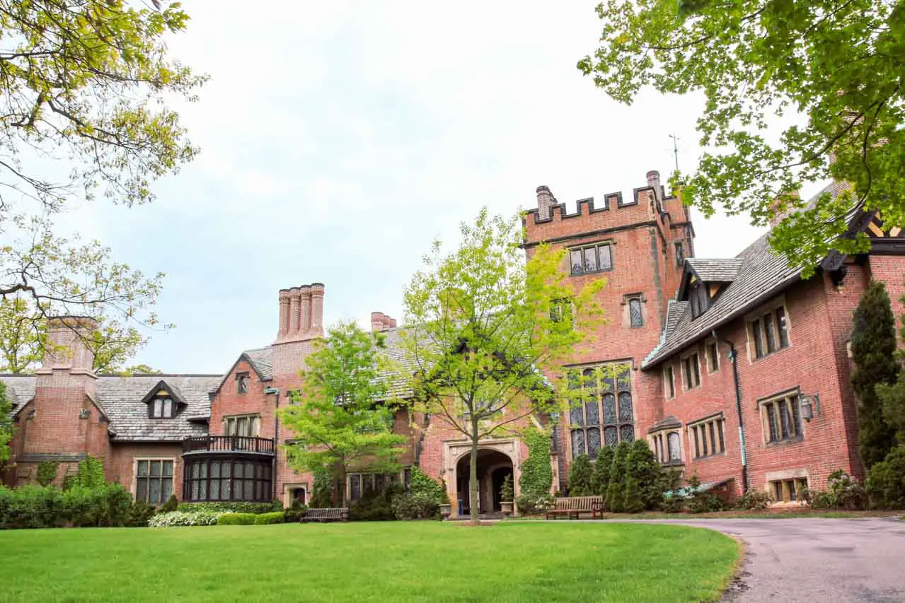 Photo of front exterior of a Tudor Revival mansion framed by trees and lawn.