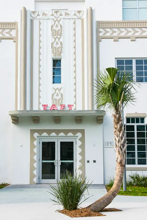 Facade of white and caramel art deco building with palm tree in foreground and illuminated sign reading "Taft"