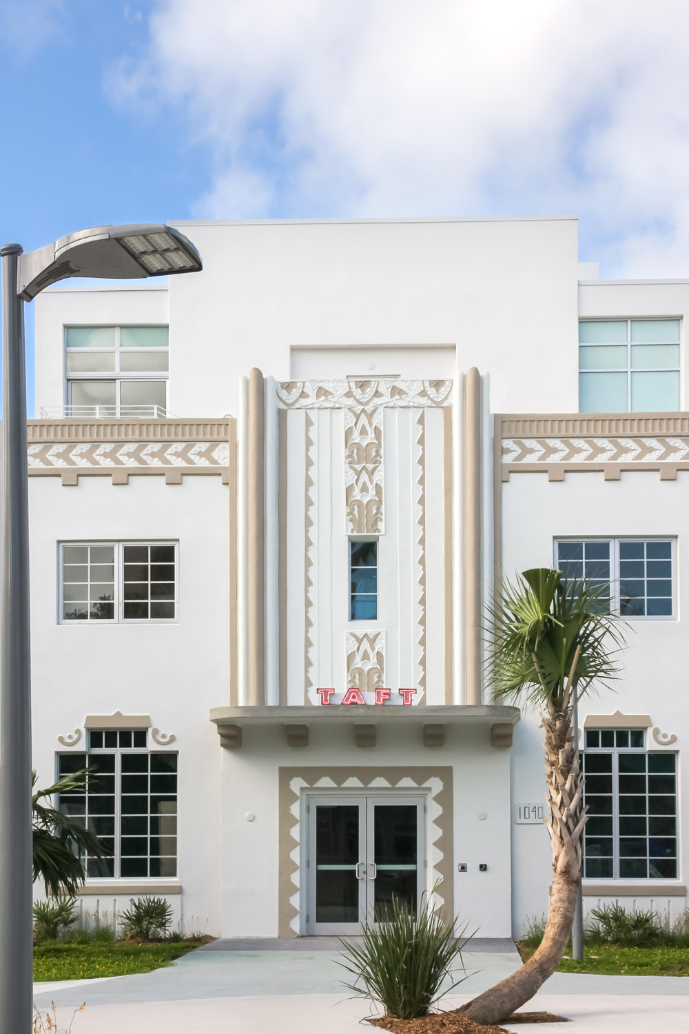Facade of white and caramel art deco building with palm tree in foreground and illuminated sign reading "Taft"