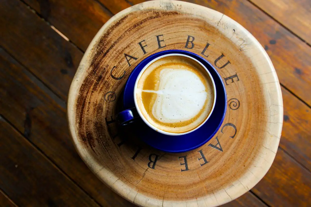 Photo of latte in blue cup and saucer resting on a wooden stool engraved with the name "Cafe Blue"