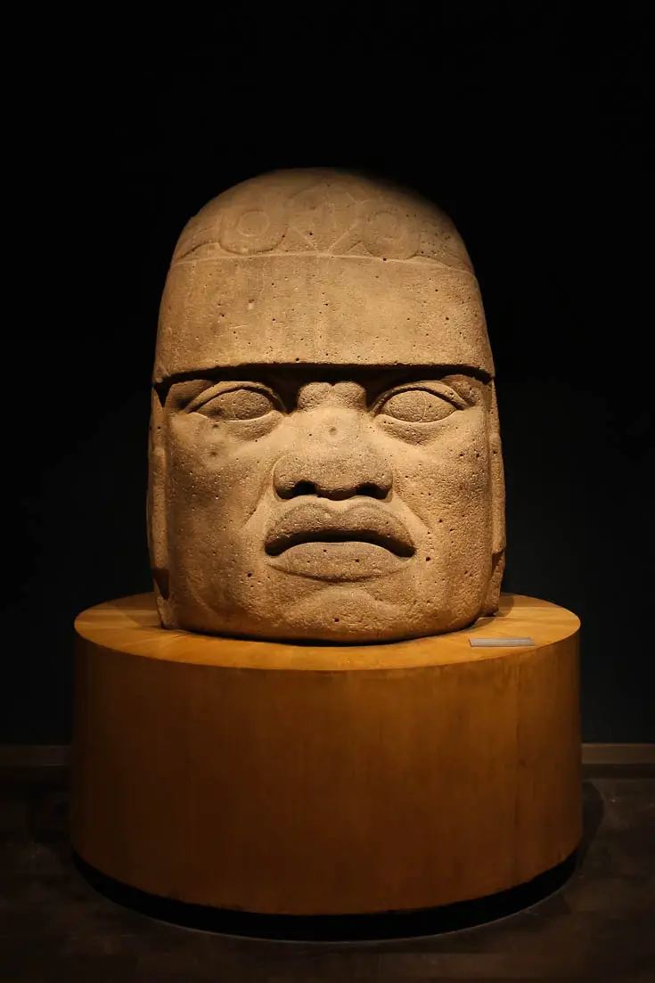 Giant head carved from basalt, sitting on a wooden plinth in a museum