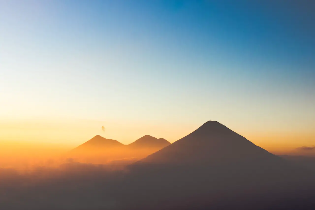Silhouette of three volcanoes against a sunset sky with the most distant one giving off a puff of smoke