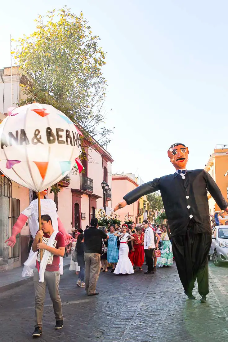 Large paper mache figure of groom and man holding giant balloon bearing names of couple at the head of a wedding recessional