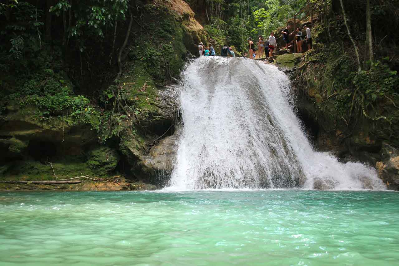 Waterfall viewed from turquoise plunge pool, with people gathering at the top of the cascade