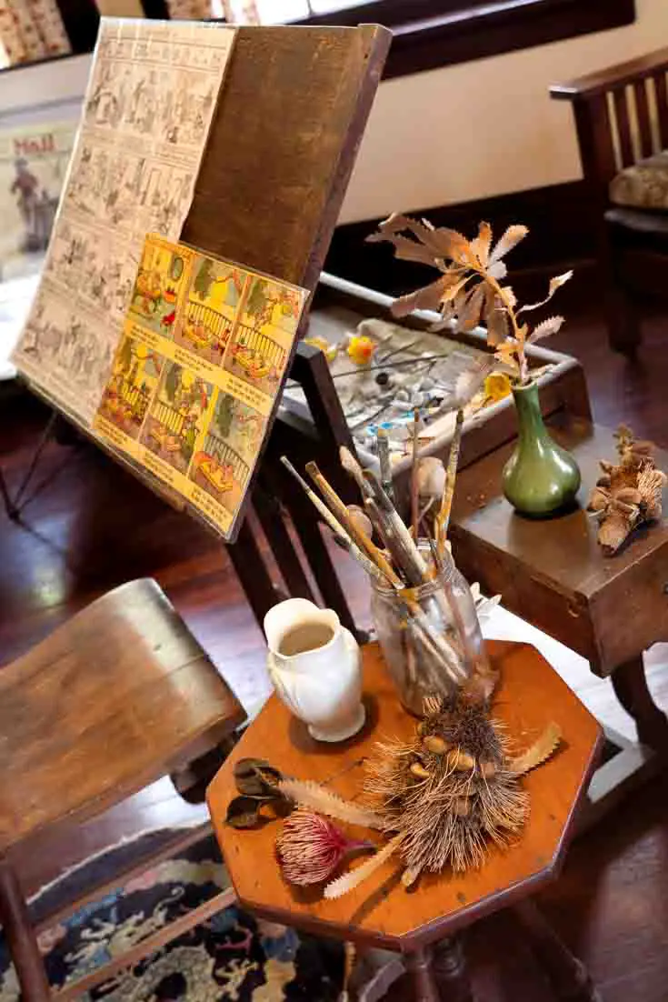 Photo of artist's easel with illustrations and a sidetable holding paintbrushes and dried native Australian plants