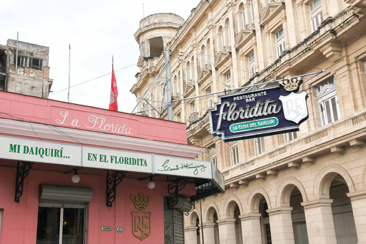 Pink exterior of Floridita with blue and green sign. Cream colonial building in background.