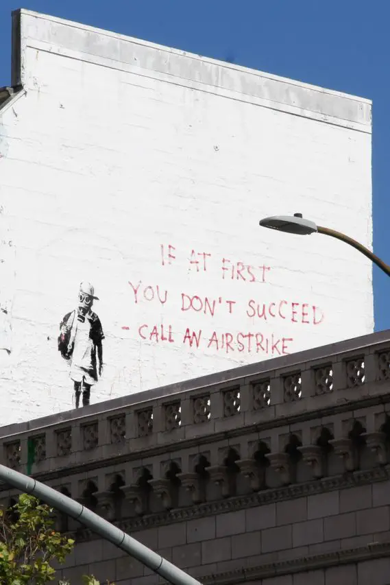 Street at painted on white wall depicting young boy in gas mask and text reading "if at first you don't succeed - call an airstrike"