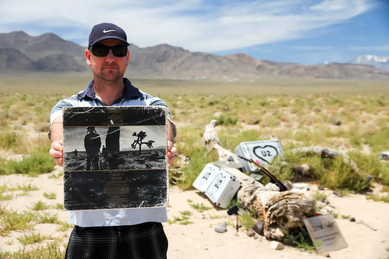 My husband holding a tattered copy of the Joshua Tree album sleeve with the falllen tree and tributes in the background