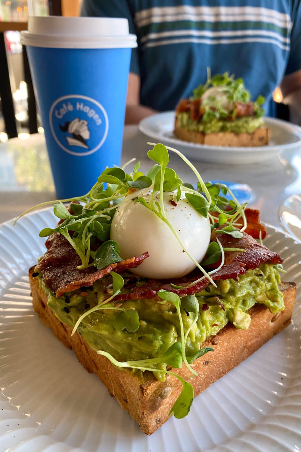 Avocado toast topped with a boiled egg and bacon, with a blue coffee cup bearing the logo of Cafe Hagen