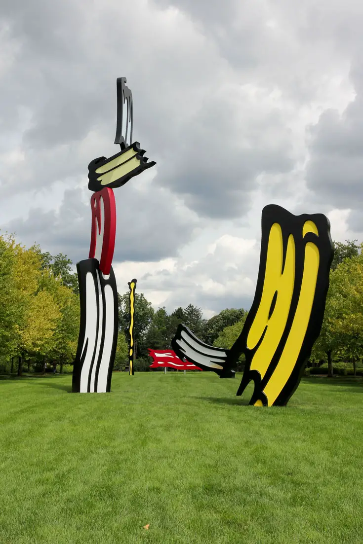 Outdoor artwork of three-dimensional paint brushstrokes on a lawn surrounded by trees and a cloudy sky