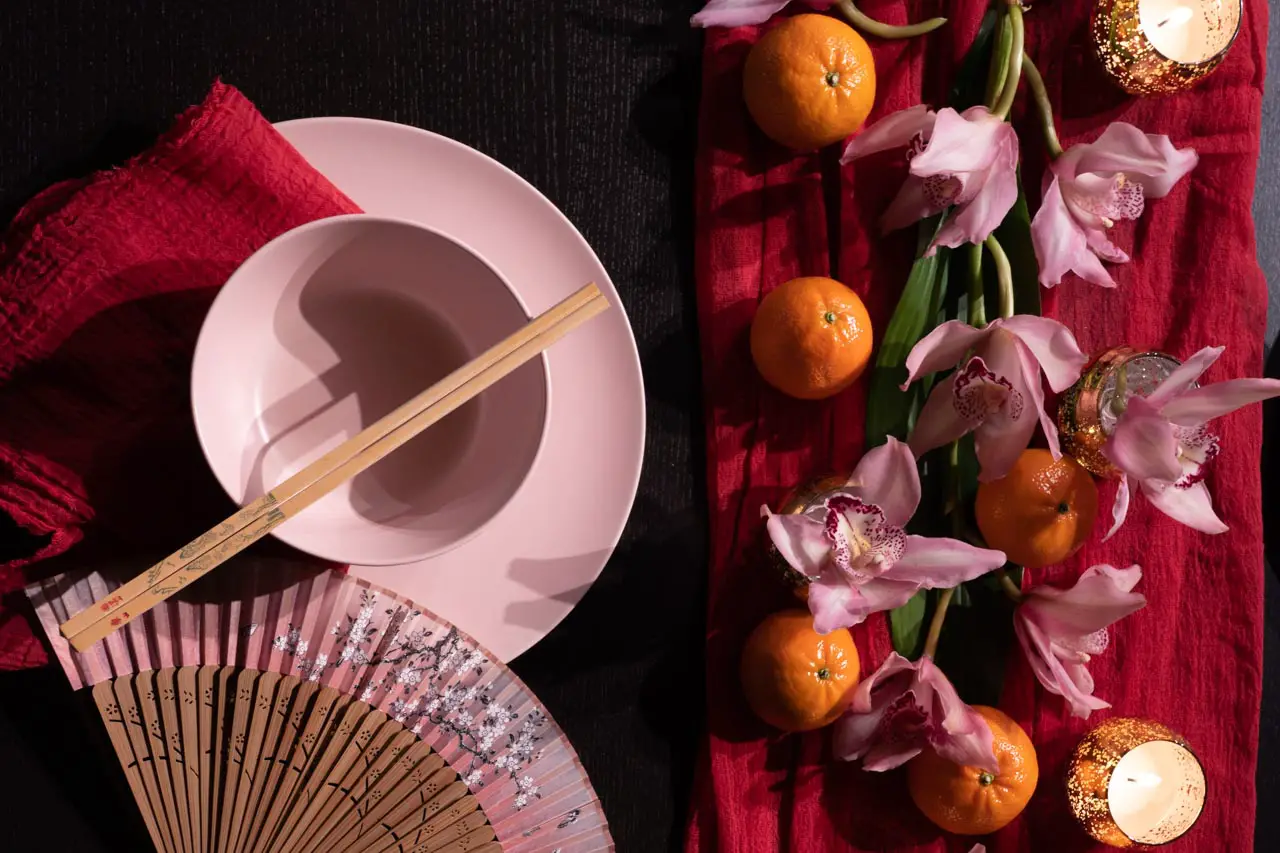 Pink bowl with chopsticks and pink folding fan on a dark table with red linen, pink orchids, mandarins and gold tealight candles.