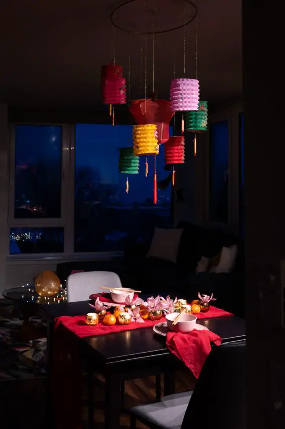 Table set for Chinese New Year dinner in red, pink and gold with colourful paper lanterns hanging above.