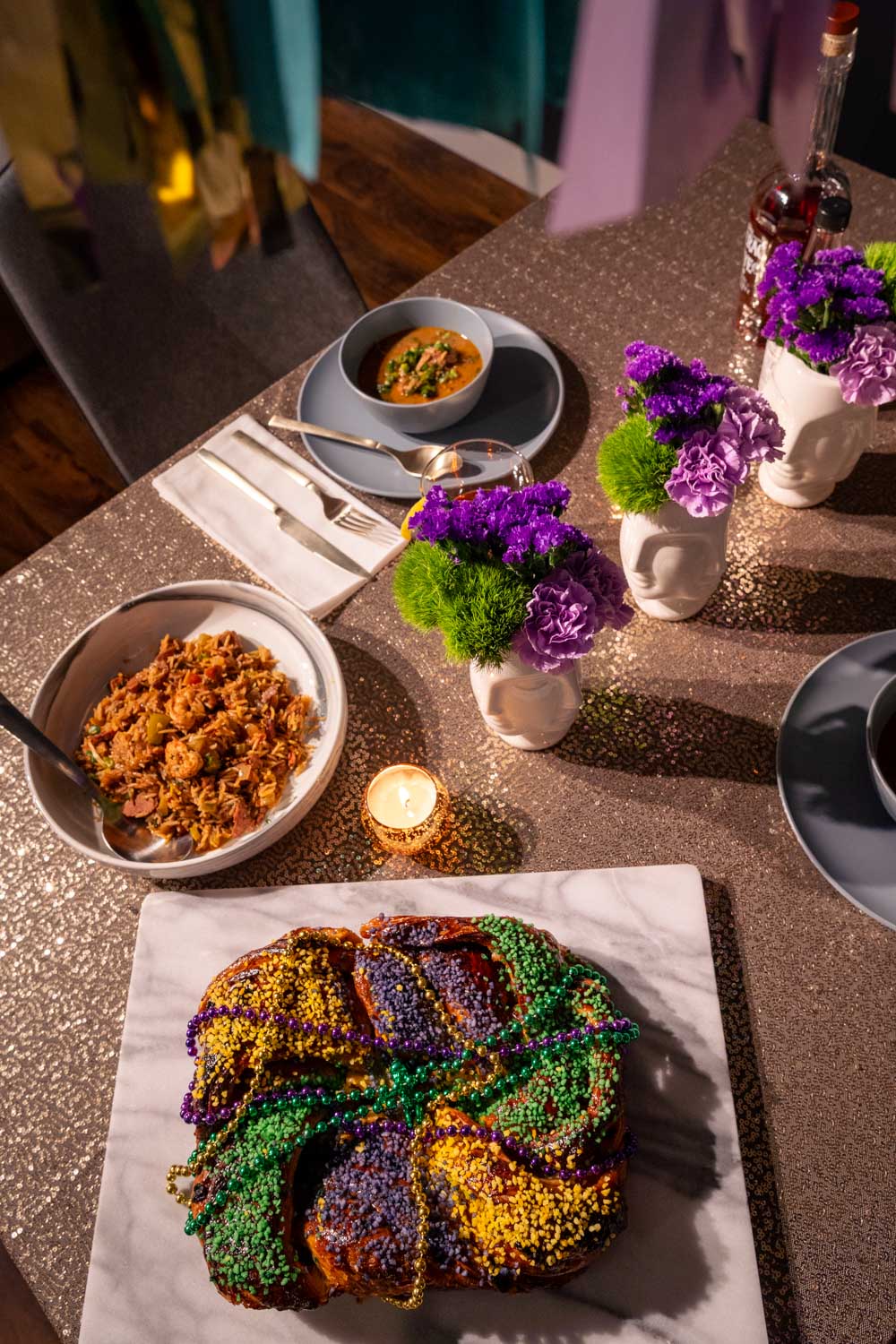 Photo of tabletop with gold sequin tablecloth, purple and green flowers, gold tealight candle holders, plates of gumbo, jambalaya and King Cake.