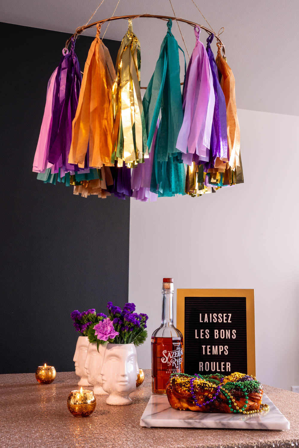Paper tassel chandelier over a table with gold sequin tablecloth, king cake, bottle of Sazerac and sign that says "Laissez les bons temps rouler"