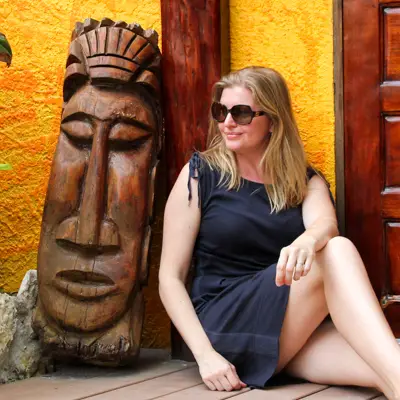 Madam ZoZo in blue dress and sunglasses sitting besides wood carving of face, against bright yellow wall