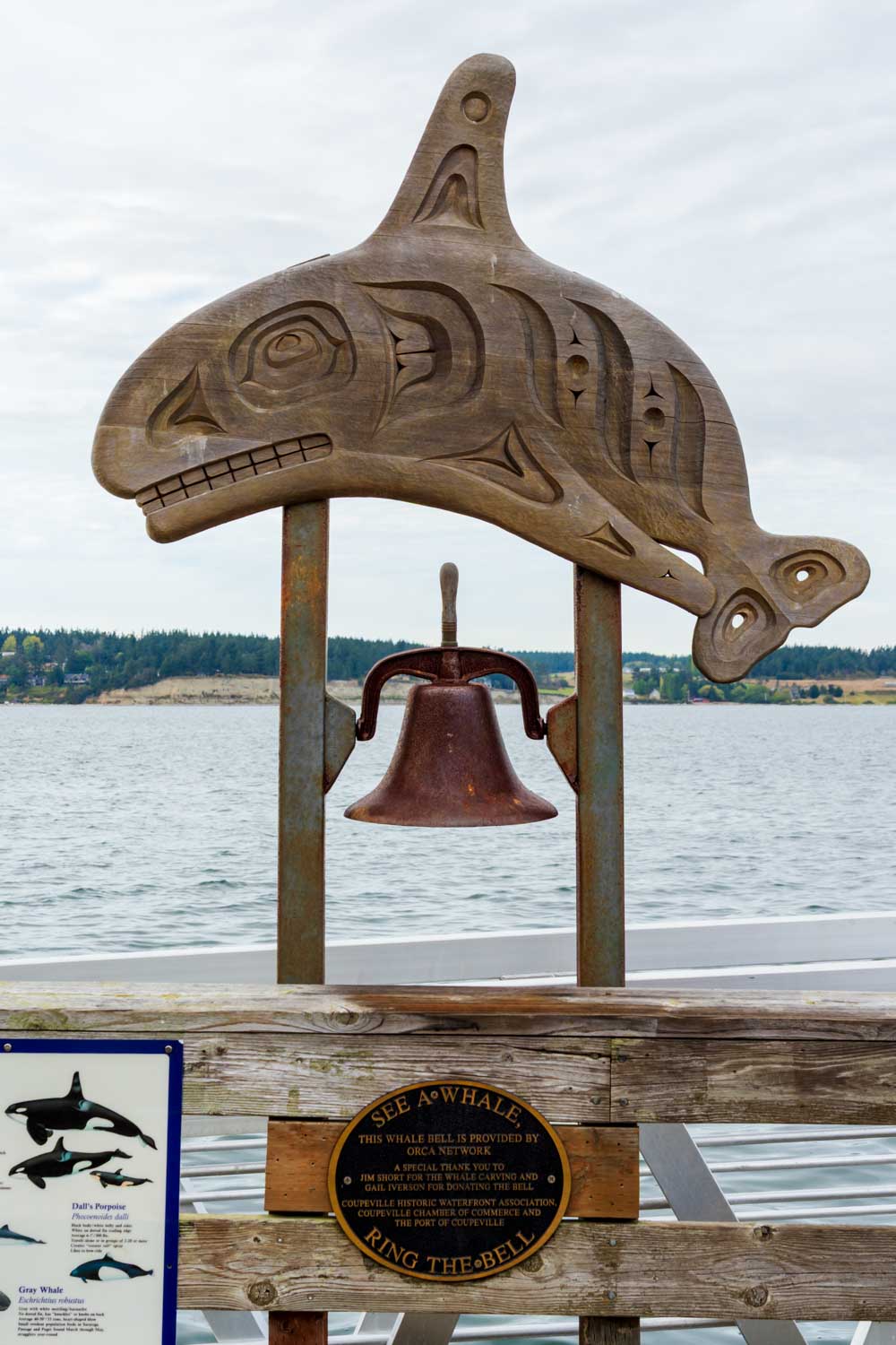 Indigenous wood carving in the shape of a whale with a lare bell hanging below, facing the water