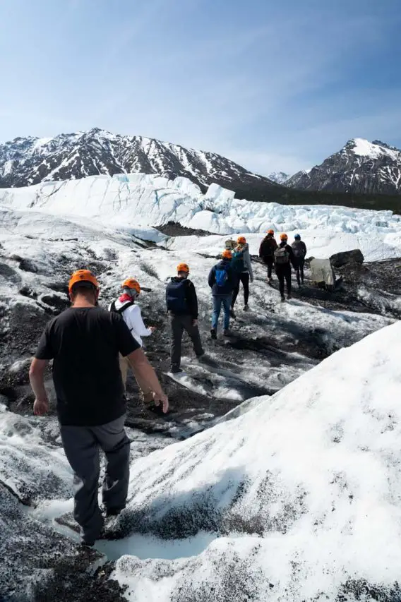 Small group of people in orange helmets, hiking in single-ile along a glacier
