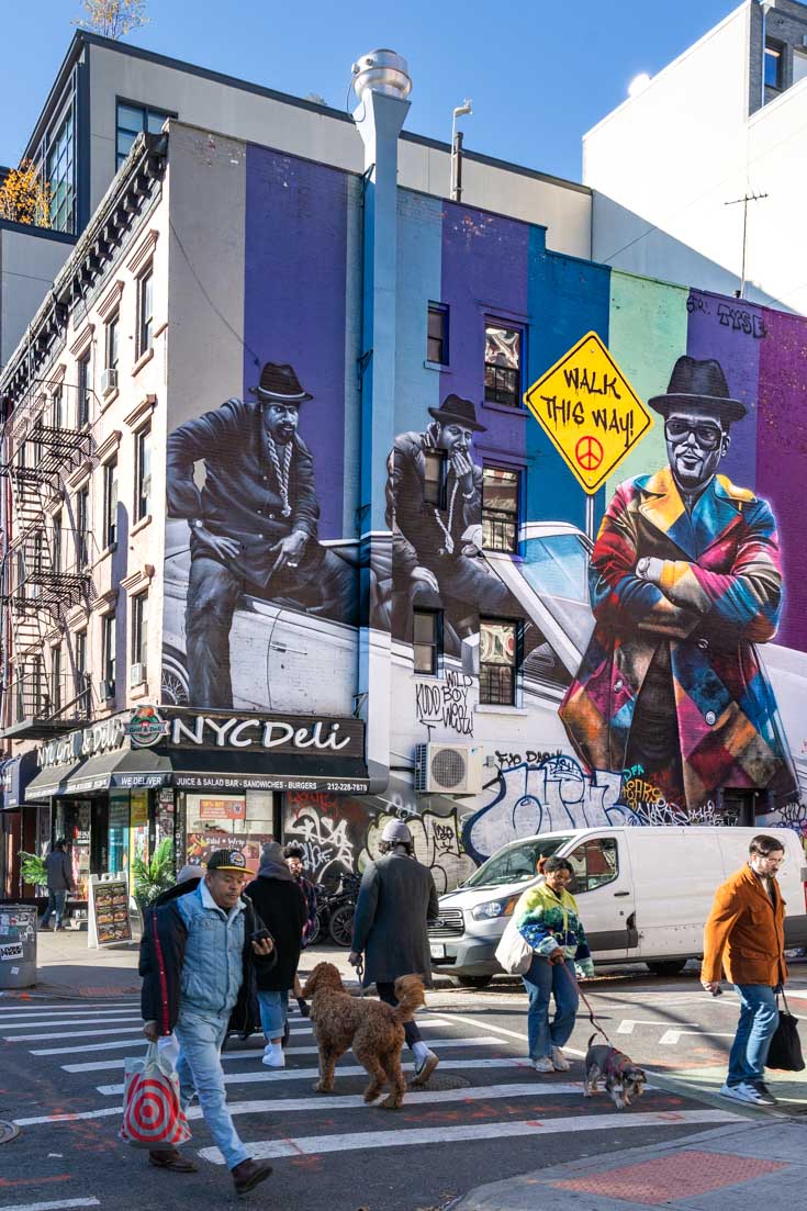 Mural of Run DMC with street sign reading "walk this way" in foreground are people crossing street