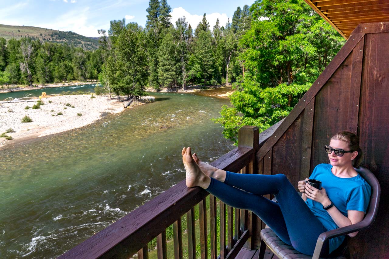 Woman holding a mug of tea, with her feet up on a balcony overlooking the confluence of two rivers on a sunny day