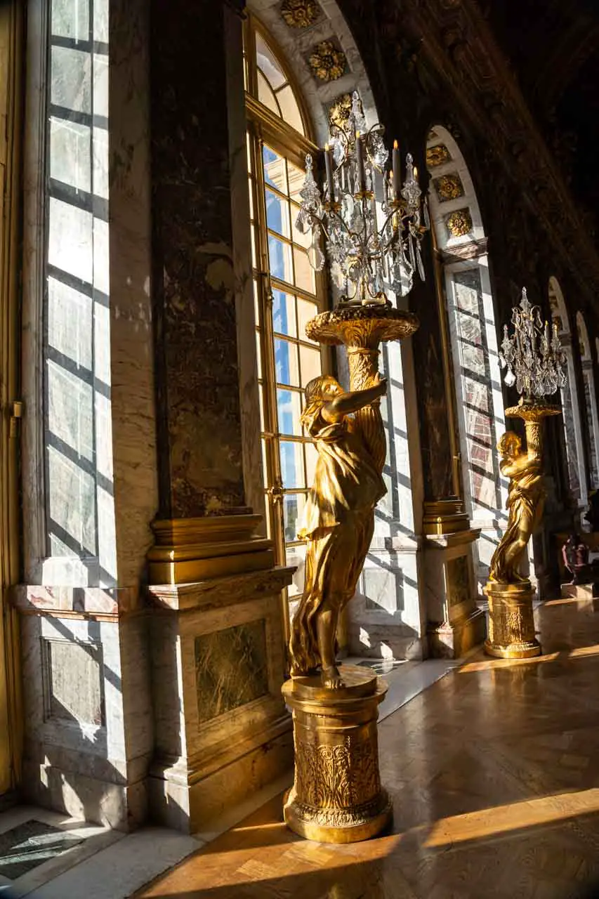 Hall of Mirrors in Versailles with light streaming through arched windows on to gold sculptures holding up crystal chandeliers