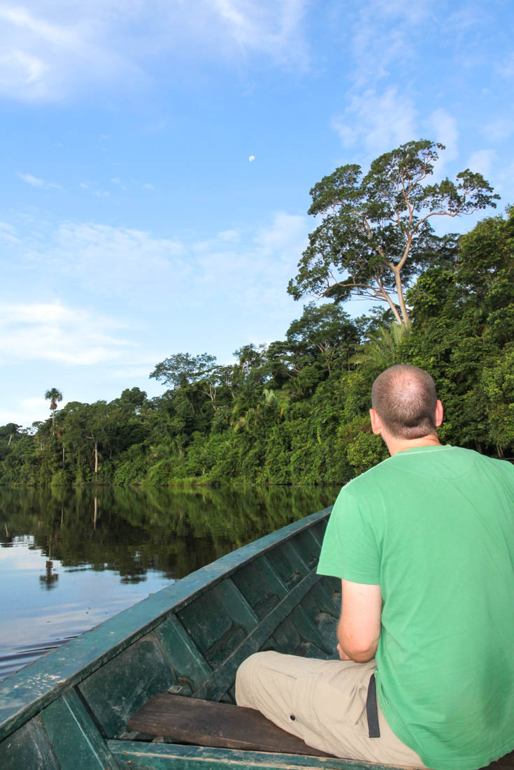 Man in green shirt at the front of a boat, flating across a still lake, the waters reflecting the tropical rainforest around the lake's perimeter.