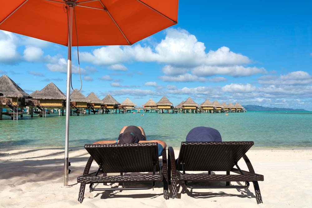 Couple on sunloungers below a red beach umbrella, looking out at tropical water and overwater bungalows