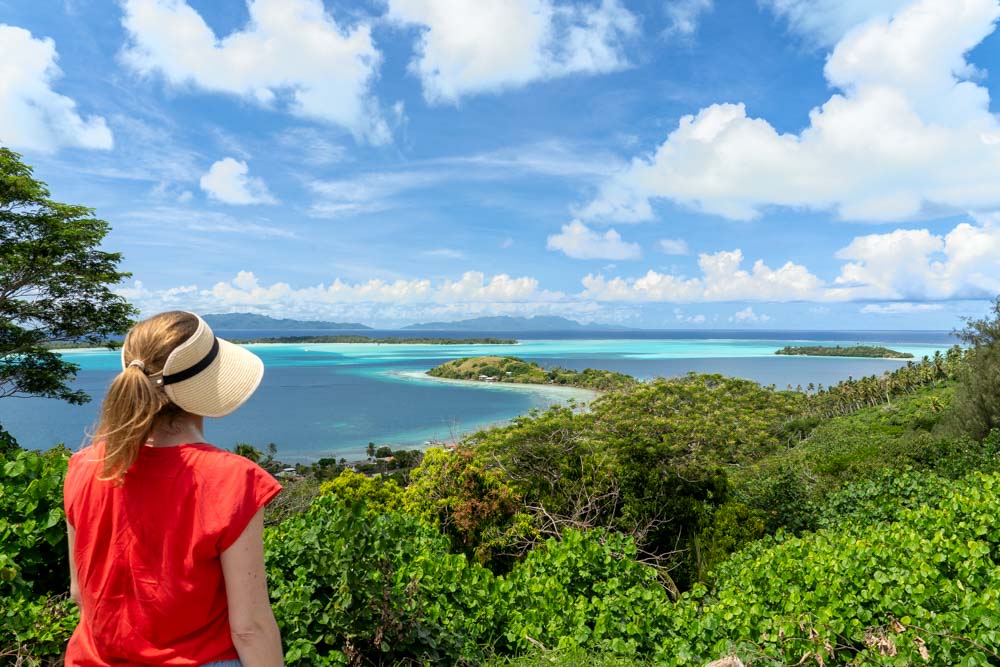 Woman in straw visor and red shirt looking out at tropical lagoon, with lush green forest-lined hillside in the foreground