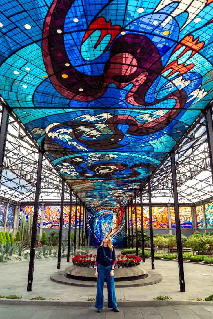Woman standing under large stained glass mural in indoor botanic garden