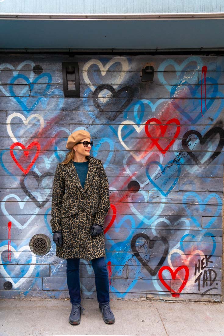 Woman in leopard print coat and beret standing infront of street art featuring blue, red and white hearts