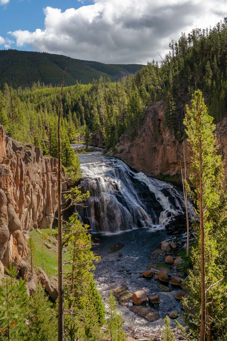 Photo of waterfall cascading into rock walled canyon from above, surrounded by forested hills