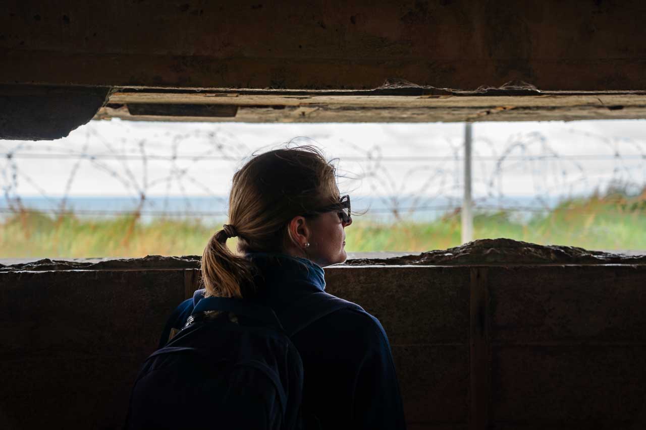 Woman looking out at barbed wire and the ocean from a dark bunker