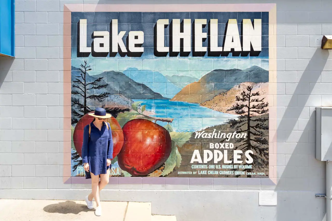 Woman in blue dress and wide brim hat standing infront of vintage style mural reading "Lake Chelan - Washington Boxed Apples"