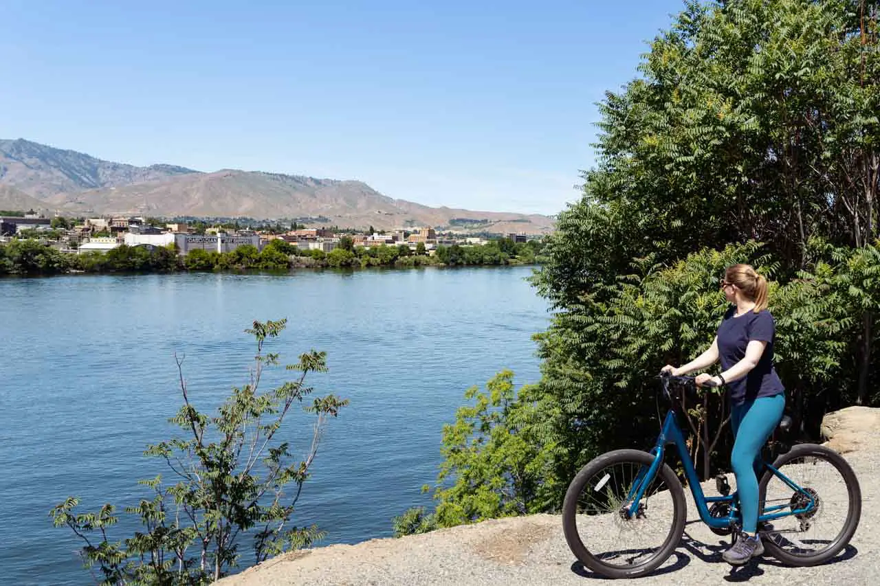 Woman on bicycle looking across wide river to rural town and mountains beyond