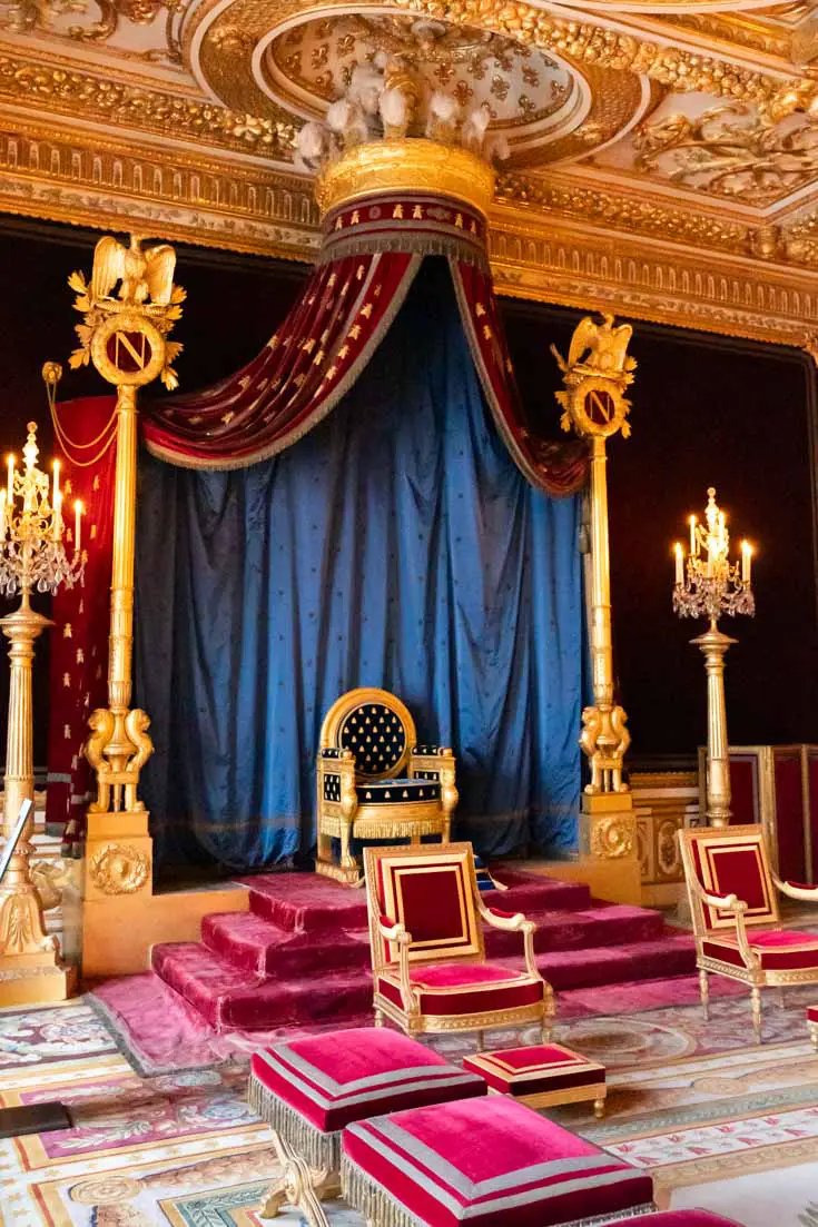 Image of small gold throne on raised platform lined with red velvet carpet and framed with royal blue canopy