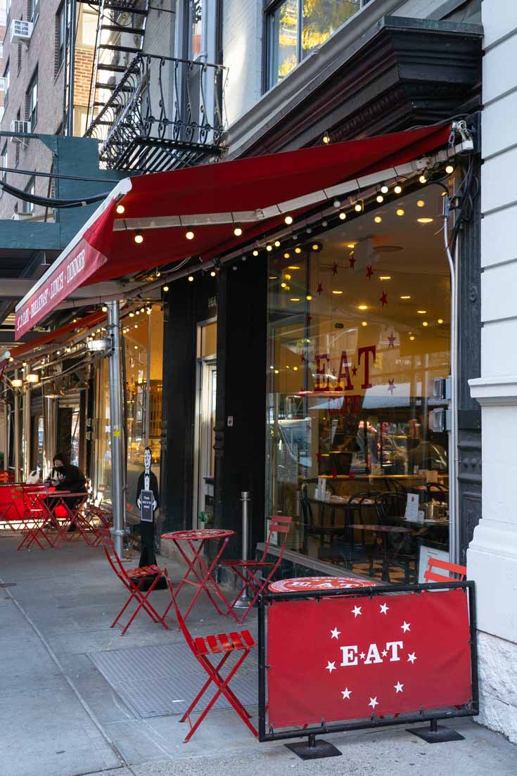 Sidewalk cafe vignette with red awning, tables and chairs, and sign reading "E.A.T"