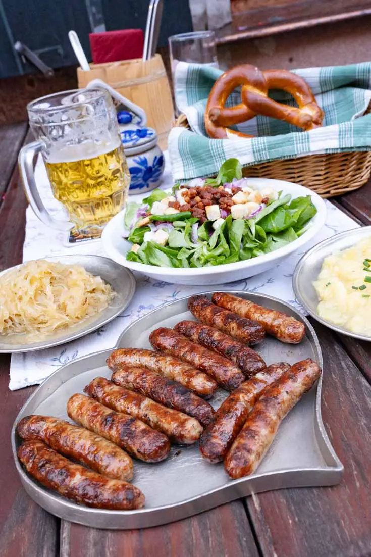 Heart-shaped metal plate of Nuremberg Bratwurst, with plates of potato salad, a pretzel and beer