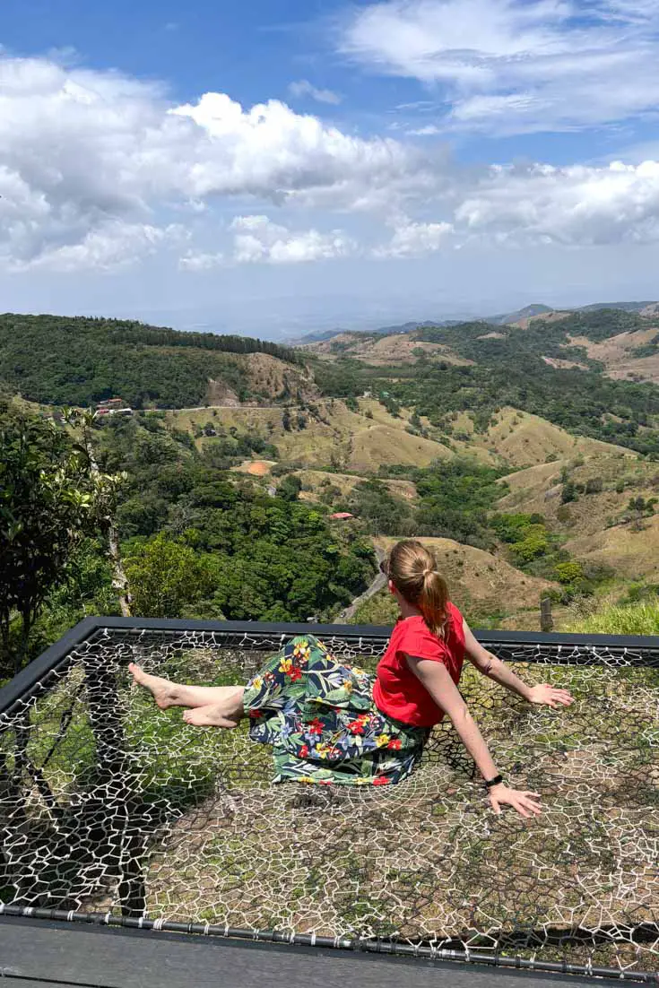 Woman in red top and tropical print skirt sitting on trampoline overlooking mountain scenery