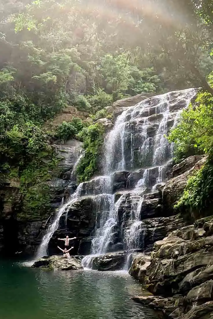 Photo of couple at the base of a large cascading waterfall in the rainforest