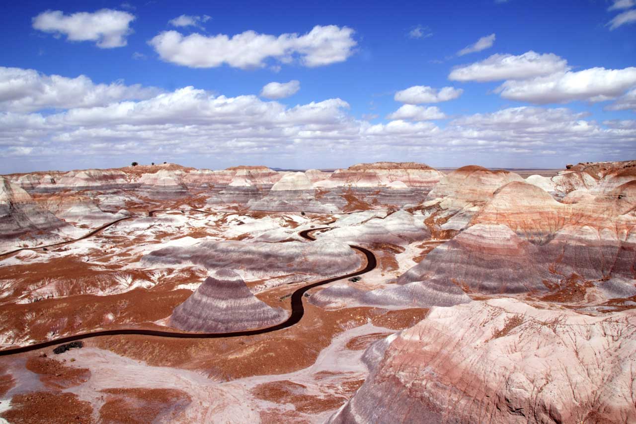 Photo overlooking road winding through colourful desert gelogy under blue, partially cloudy sky