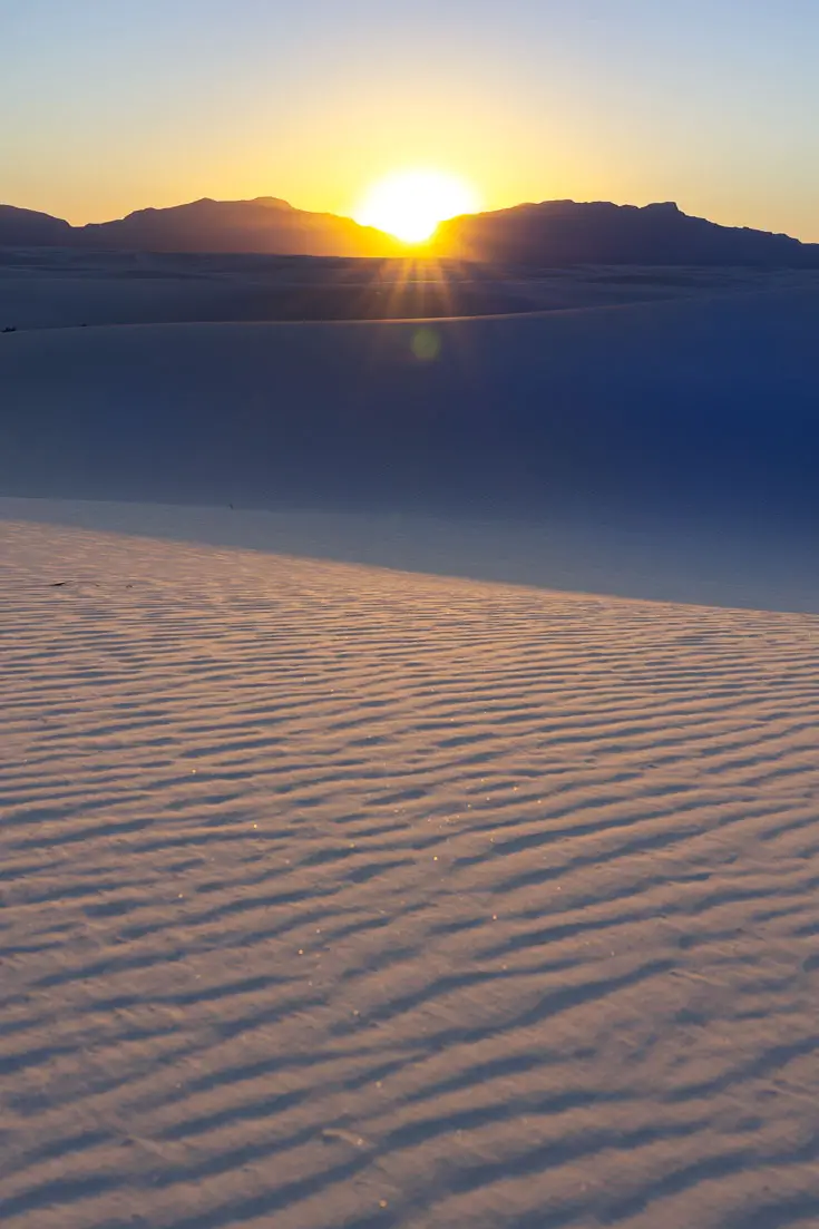 Sun just dipping below distant mountains with rippled sand dunes in foreground