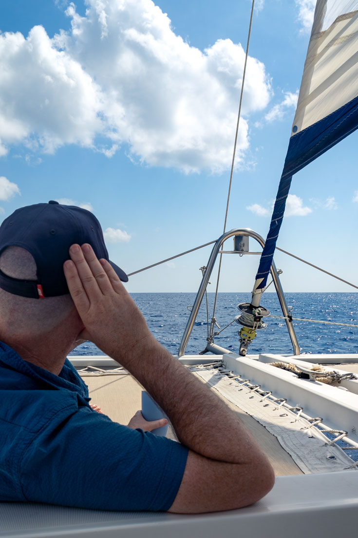 Man relaxing on deck of a catamaran with a drink, looking out to blue sky and ocean