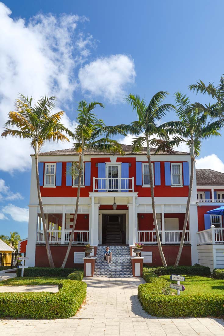 Entrance to a red, blue and white, colonial mansion framed by tall palm trees and hedges