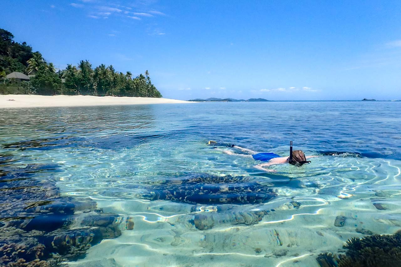 Woman in blue swimsuit, snorkelling in crystal-clear water off white-sand beach